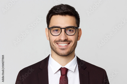 Close up headshot of young businessman wearing glasses and smart casual suit, smiling at camera, isolated on gray background