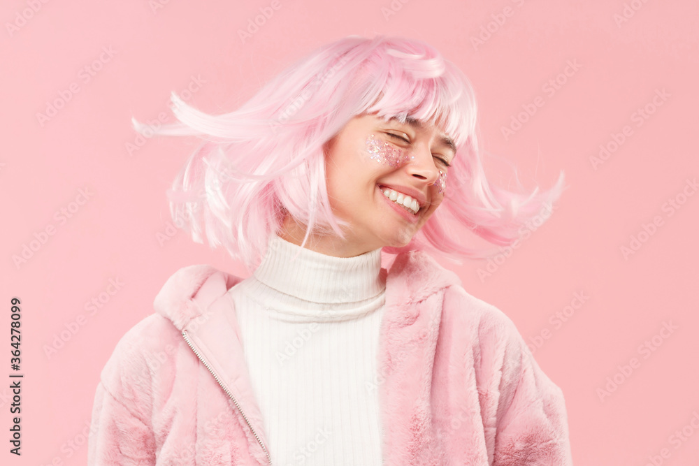 Young happy girl with closed eyes in fur coat, dancing and moving head so her hair is flying, isolated on pink background
