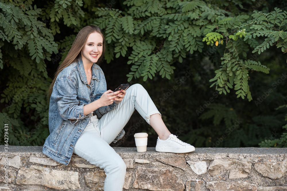 blonde girl with smartphone looking at camera. Girl dressed in pants and denim jacket sitting on a stone wall blonde girl with smartphone looking at camera. Girl dressed in pants and denim jacket sitting on a stone wall