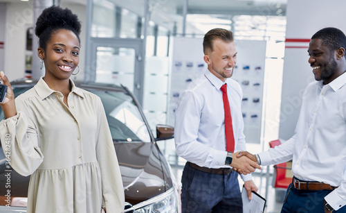 portrait of young black lady with keys of new car in hands, she looks at camera and smile. two men shake hands in the background. in cars showroom