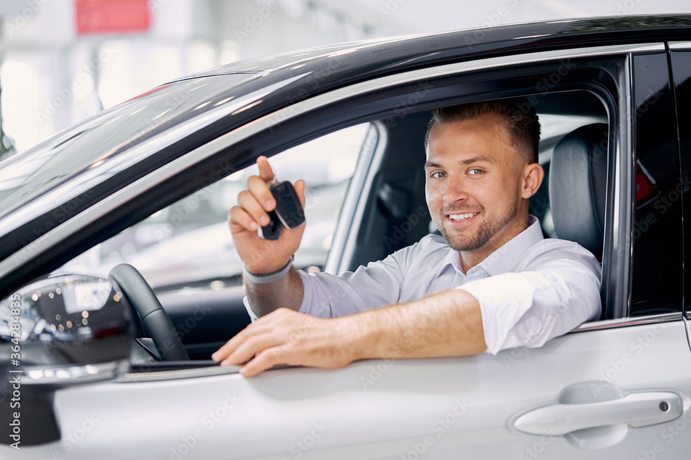 young caucasian man is happy to get keys of auto, he made purchase in modern car showroom