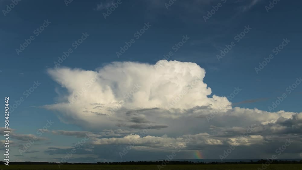 Time lapse of a large billowing thundercloud with a rainbow below the cloud. A crop of bright yellow canola is in the foreground and sunbeams flicker across the field. Time lapse of a large billowing thundercloud with a rainbow below the cloud. A crop of bright yellow canola is in the foreground and sunbeams flicker across the field.