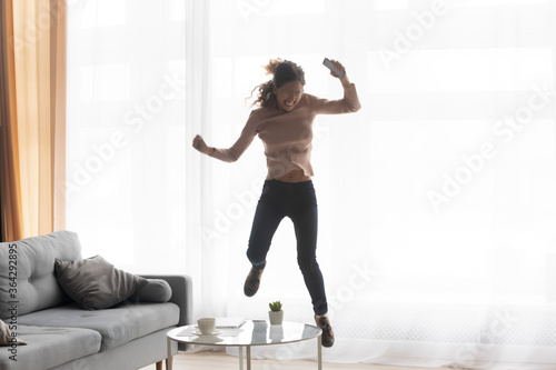 Fotografie, Tablou Excited young Caucasian woman have fun jumping dancing celebrate win in living r