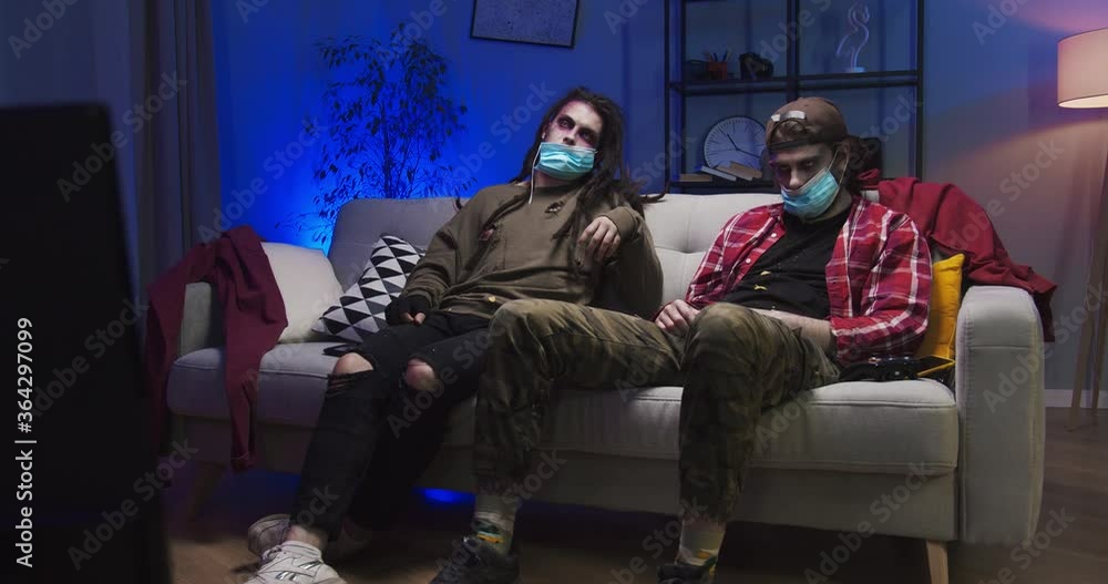 Scary male zombies watching TV at home during pandemic period, self-isolation. Frightening infected dead men sitting on sofa in masks. Halloween, costume party, supernatural concept. Trick or treat. Scary male zombies watching TV at home during pandemic period, self-isolation. Frightening infected dead men sitting on sofa in masks. Halloween, costume party, supernatural concept. Trick or treat.