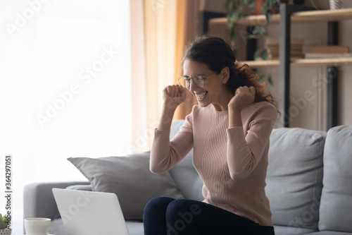 Excited millennial girl in glasses sit on sofa at home triumph reading good news promotion email on laptop, overjoyed young Caucasian woman look at computer screen feel euphoric win lottery online