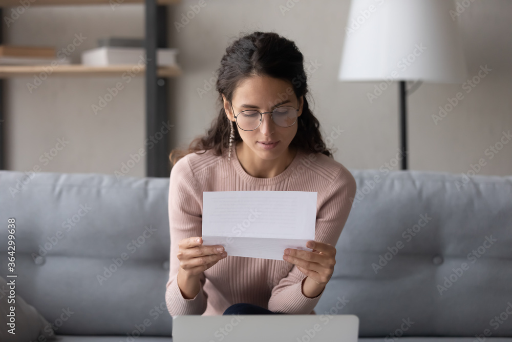 Serious young Caucasian woman in glasses sit on couch at home reading paper postal letter, concentrated millennial female in eyewear consider paperwork correspondence, work news offer in post notice Serious young Caucasian woman in glasses sit on couch at home reading paper postal letter, concentrated millennial female in eyewear consider paperwork correspondence, work news offer in post notice