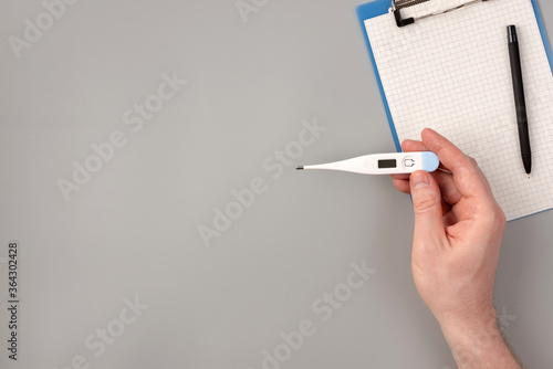 Thermometer in hand, notepad and pen on gray background, health care concept and prevent the spread of pandemic Covid-19, coronavirus