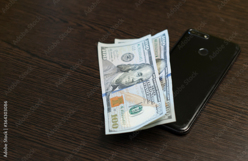 One hundred dollar bills and smartphone lie on a wooden table. Mobile banking concept with copy space. Close-up. One hundred dollar bills and smartphone lie on a wooden table. Mobile banking concept with copy space. Close-up.