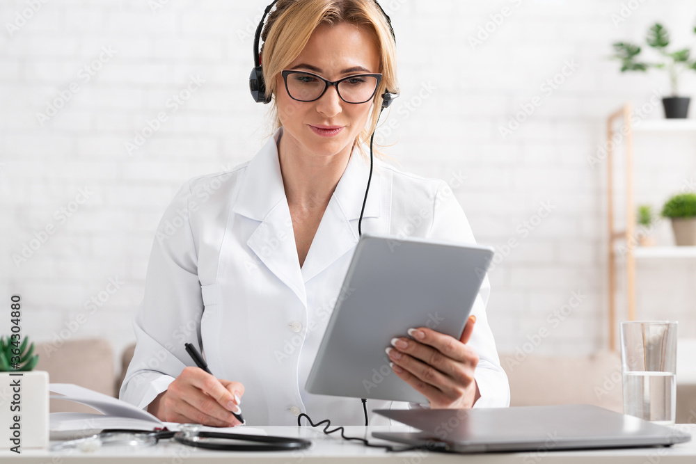Online work with patients. Busy doctor holds tablet in hands and makes notes in notebook Online work with patients. Busy doctor holds tablet in hands and makes notes in notebook