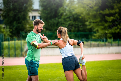 Male personal trainer assisting woman leg and shoulder stretching and talking before training in park.