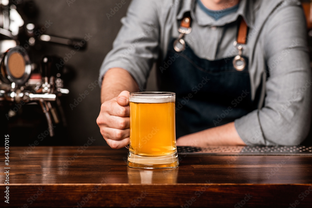 Friendly bartender. Man in apron puts a mug of light beer on wooden counter in interior
