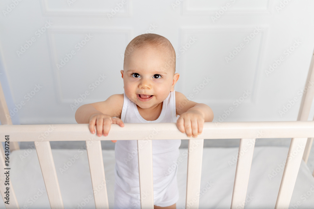 portrait of a smiling baby boy 8 months old standing in a crib in a children's room in white clothes and looking at the camera, morning baby, baby products concept portrait of a smiling baby boy 8 months old standing in a crib in a children's room in white clothes and looking at the camera, morning baby, baby products concept