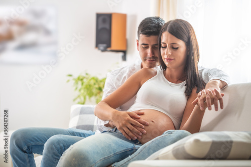 Happy couple expecting a baby sits on a couch, with both holding hands on the belly