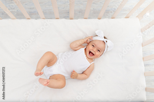 baby girl 6 months old lies in a crib in the nursery with white clothes on her back and laughs, looks at the camera, baby's morning, baby products concept