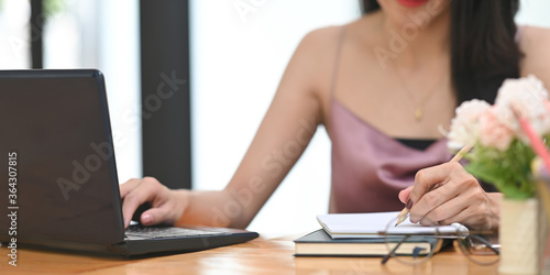 Cropped image woman is taking notes and using a computer laptop at the wooden working desk. © Prathankarnpap