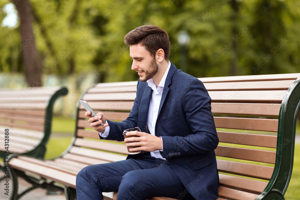 Handsome office employee having coffee to go and browsing web via smartphone on bench at park Handsome office employee having coffee to go and browsing web via smartphone on bench at park