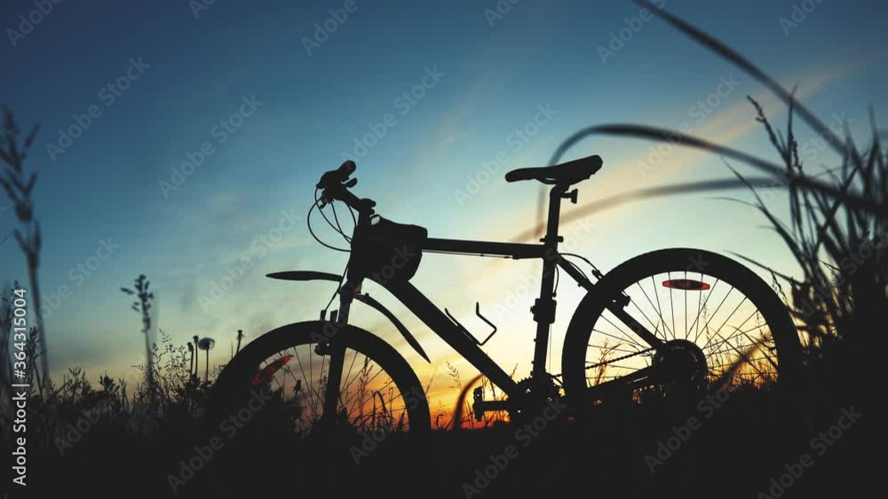 Traveling by bike. A silhouette of a bicycle stands in a field among grass on a sunset background. Soft golden fill light Traveling by bike. A silhouette of a bicycle stands in a field among grass on a sunset background. Soft golden fill light