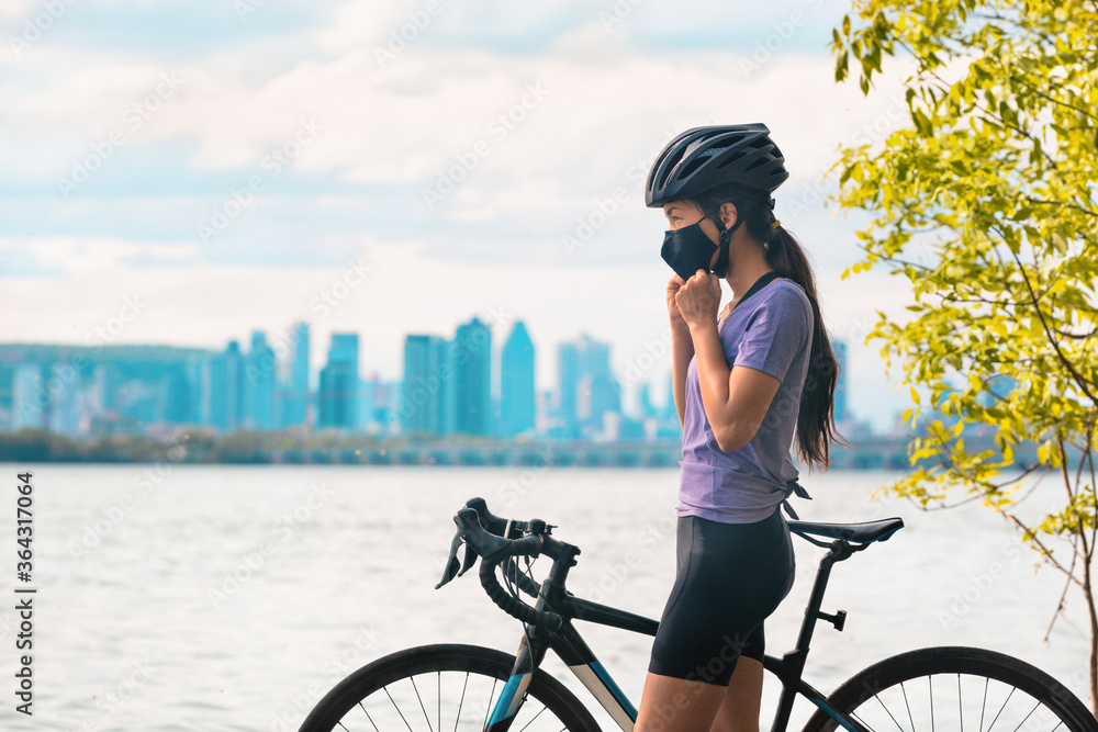 Fototapeta premium Noszenie maski covid-19 podczas jazdy na rowerze. Sportowa rowerzysta kobieta na rowerze zakładająca maskę na twarz w celu zapobiegania Covid-19 podczas letnich ćwiczeń rekreacyjnych na świeżym powietrzu. Fitness na zewnątrz.