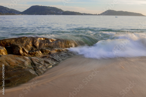 A view of a sandy beach with a large stone on the shore about which waves are beating, long exposure.