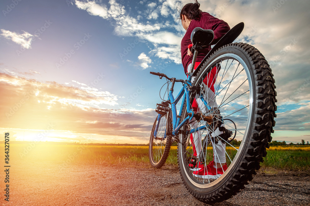 Naklejka premium Beautiful girl in a red sports suit on a bicycle on a sunset background. The concept of a healthy lifestyle, sports training, cardio load. Copy space.