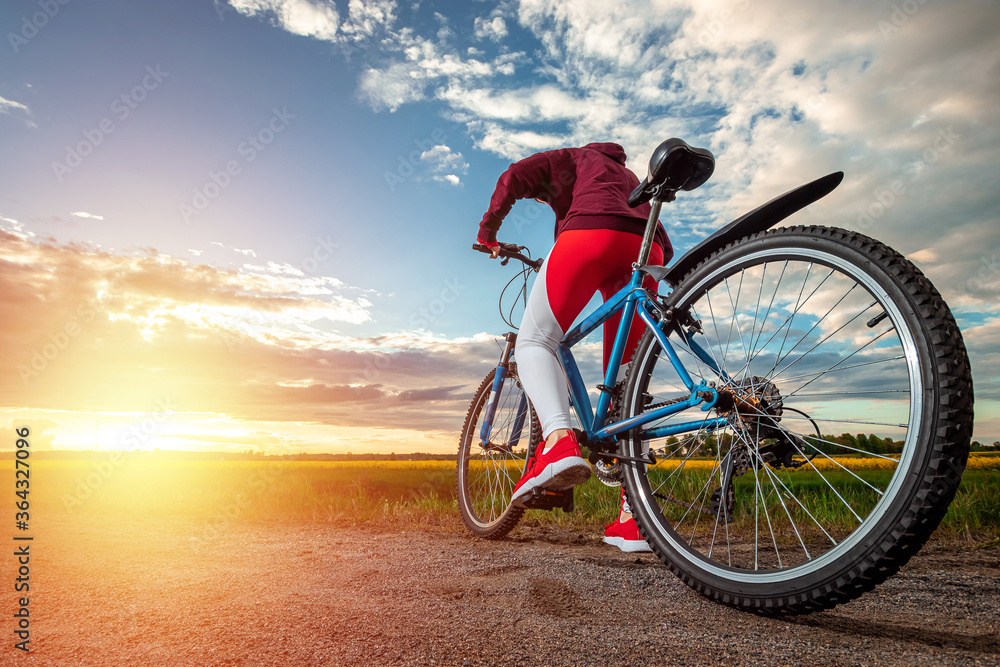 Fototapeta premium Beautiful girl in a red sports suit on a bicycle on a sunset background. The concept of a healthy lifestyle, sports training, cardio load. Copy space.