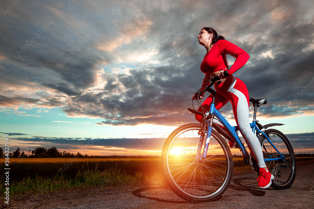 Fototapeta premium Sports training on a bicycle. Beautiful girl in a sports suit on a sunset background. The concept of a healthy lifestyle, cardio training, physical activity. Copy space.