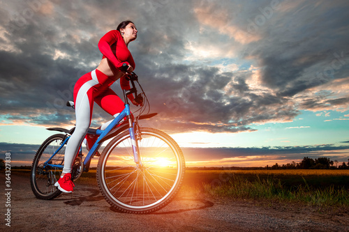 Beautiful girl in a red sports suit on a bicycle on a sunset background. The concept of a healthy lifestyle, sports training, cardio load. Copy space.