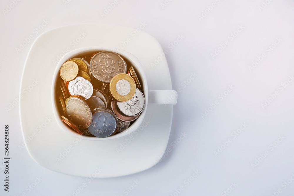 White cup of coffee with different coins on a white background. Concept of finance and business. Copy space. White cup of coffee with different coins on a white background. Concept of finance and business. Copy space.