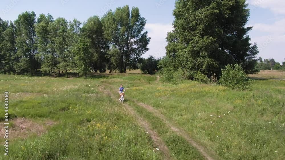 Beautiful young woman cycling in the green field towards the camera. Green field full of flowers and grass with a path surrounded by big trees. Aerial drone footage. Beautiful young woman cycling in the green field towards the camera. Green field full of flowers and grass with a path surrounded by big trees. Aerial drone footage.