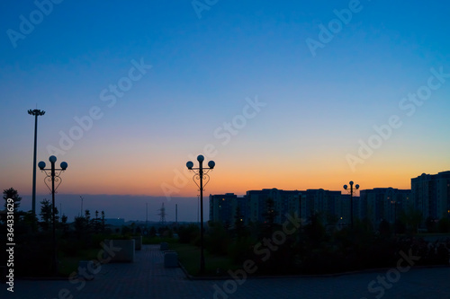 Dawn in the city. The city before dawn. Silhouettes of lanterns. The sun rises. Bright streak in the sky. Dawn sky over tall buildings. Dark square before dawn. © Эльвира Турсынбаева