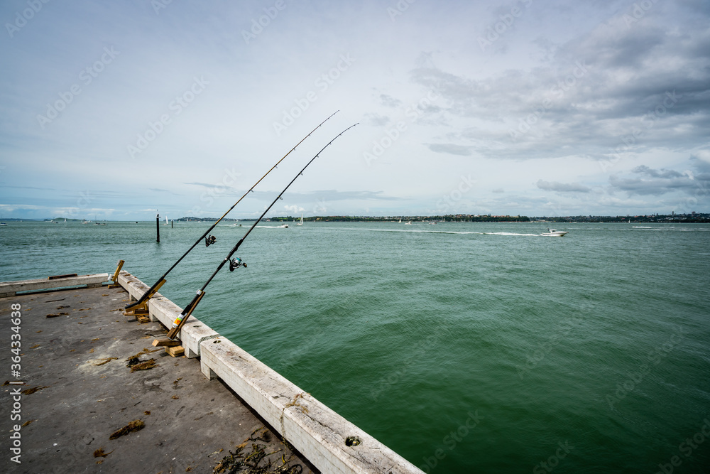 Auckland, North Island New Zealand – circa 2018: Two un-manned fishing rods set up on a pier, waiting to catch a fish. Lifestyle use. 