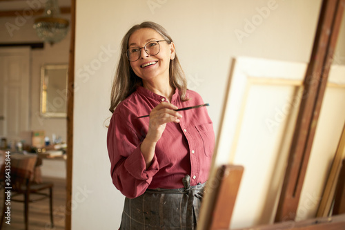 Attractive cheerful female pensioner in apron and eyeglasses imroving artistic skills, standing in studio next to easel, holding paintbrush during art workshop. People, leisure and hobby concept