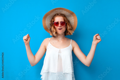 girl in a sun hat and glasses rejoices victory on a blue isolated background, joyful woman shows a win gesture in summer clothes, summer concept