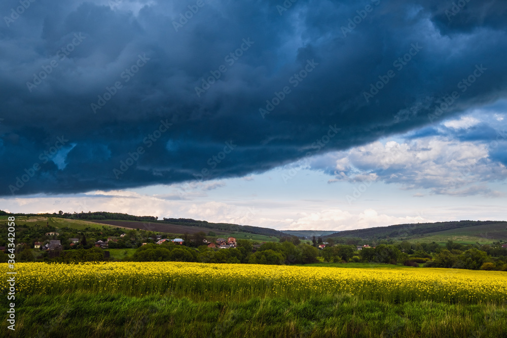 Dramatic thunderstorm cloudy sky over spring yellow flowering rapeseed fields and countryside farmlands hills. Natural weather, climate, countryside beauty concept scene.