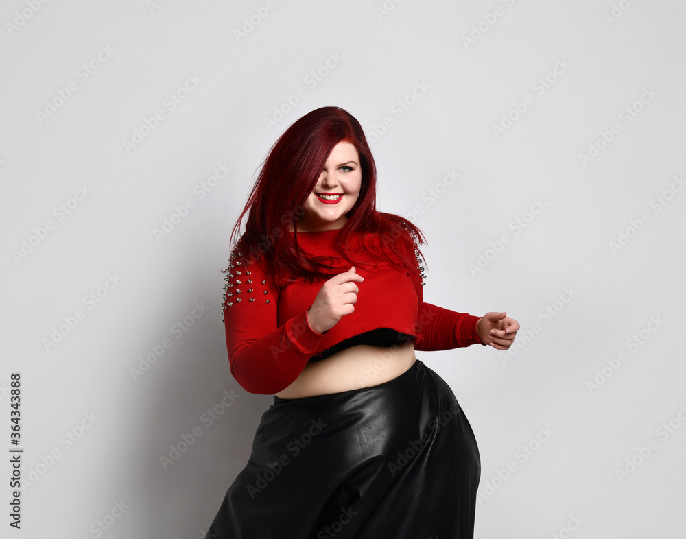 Overweight redhead woman in red spiked top, black bra and leather skirt. Dancing and smiling, posing isolated on white background Overweight redhead woman in red spiked top, black bra and leather skirt. Dancing and smiling, posing isolated on white background