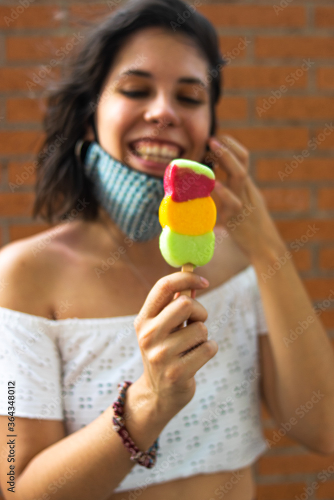 Photo of young and attractive woman wearing a reusable face mask while eating a fruit ice cream in the street with summer clothes. Photo of young and attractive woman wearing a reusable face mask while eating a fruit ice cream in the street with summer clothes.