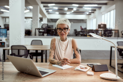 Stylish business woman. Young, positive tattooed business lady in eyewear looking at camera and smiling while working in the modern office