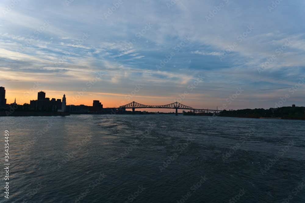 A skyline view a steel bridge across a river at the time between sunset and twilight. Jacques Cartier Bridge is across Saint Laurent river. Left is Clock Tower. Right is Saint Helen's Island.