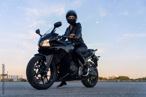Valokuva Attractive girl with long hair in black leather jacket and pants on outdoors parking with stylish sports motorcycle at sunset