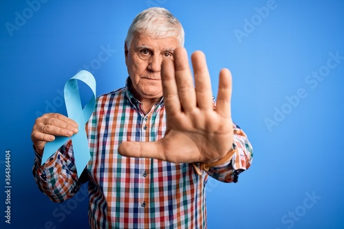 Senior handsome hoary man holding blue cancer ribbon symbol over isolated background with open hand doing stop sign with serious and confident expression, defense gesture