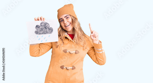 Young caucasian woman wearing winter clothes holding rain draw surprised with an idea or question pointing finger with happy face  number one