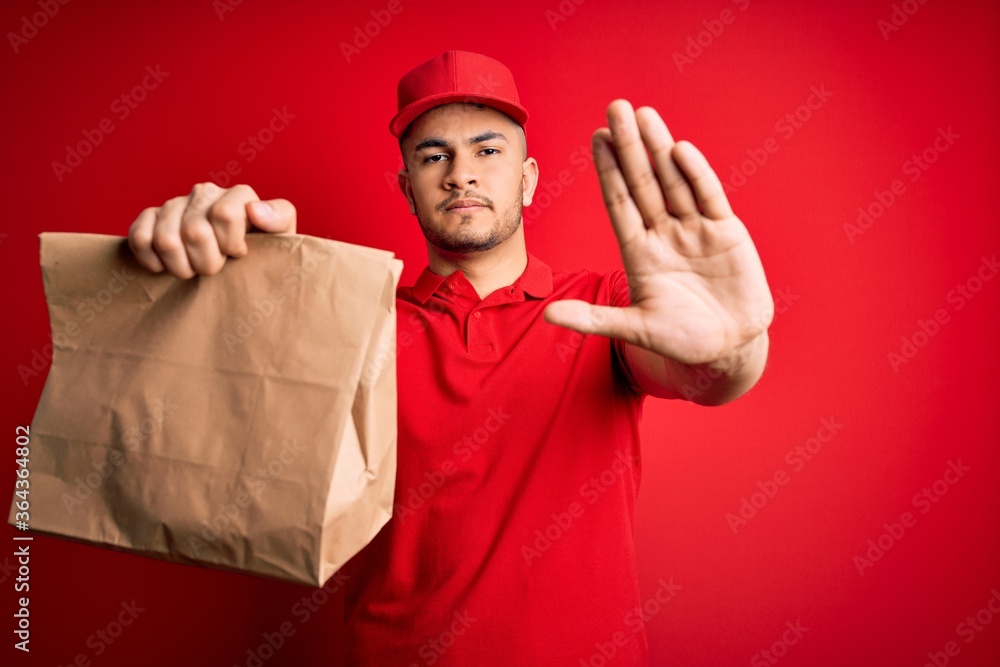 Young handsome delivery man holding paper bag with takeaway food over red background with open hand doing stop sign with serious and confident expression, defense gesture Young handsome delivery man holding paper bag with takeaway food over red background with open hand doing stop sign with serious and confident expression, defense gesture