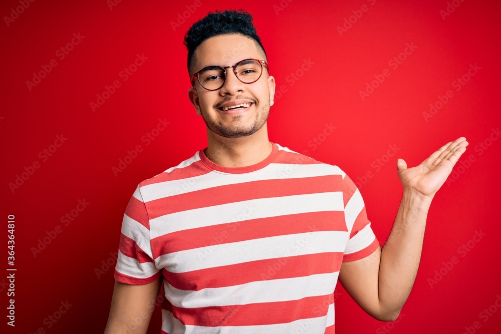 Young handsome man wearing casual striped t-shirt and glasses over isolated red background smiling cheerful presenting and pointing with palm of hand looking at the camera. Young handsome man wearing casual striped t-shirt and glasses over isolated red background smiling cheerful presenting and pointing with palm of hand looking at the camera.