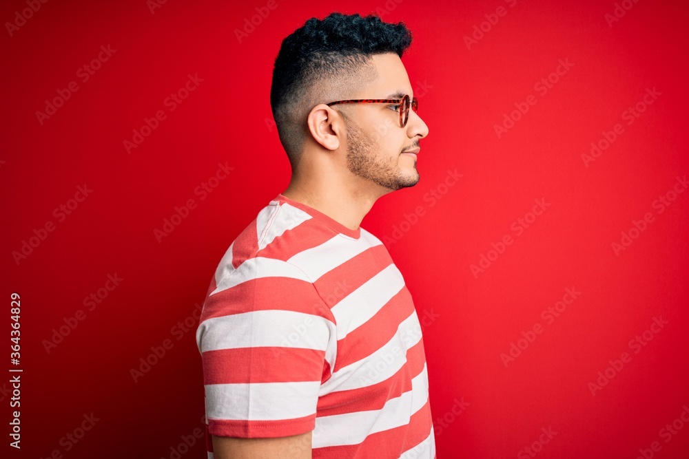 Young handsome man wearing casual striped t-shirt and glasses over isolated red background looking to side, relax profile pose with natural face and confident smile. Young handsome man wearing casual striped t-shirt and glasses over isolated red background looking to side, relax profile pose with natural face and confident smile.