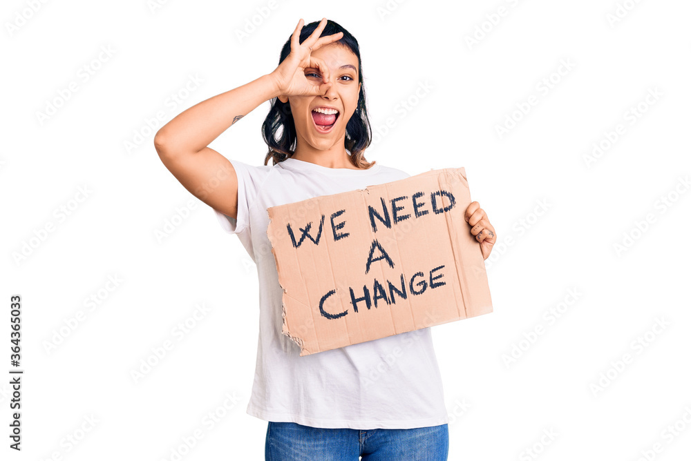 Young woman holding we need a change banner smiling happy doing ok sign with hand on eye looking through fingers Young woman holding we need a change banner smiling happy doing ok sign with hand on eye looking through fingers