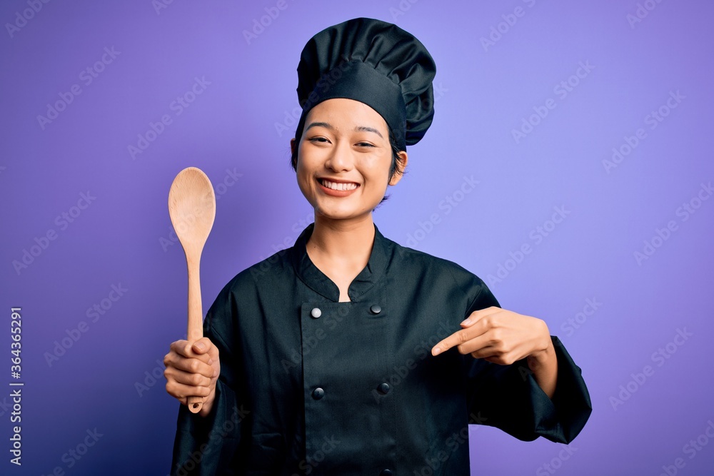 Fototapeta premium Young beautiful chinese chef woman wearing cooker uniform and hat holding wooden spoon with surprise face pointing finger to himself