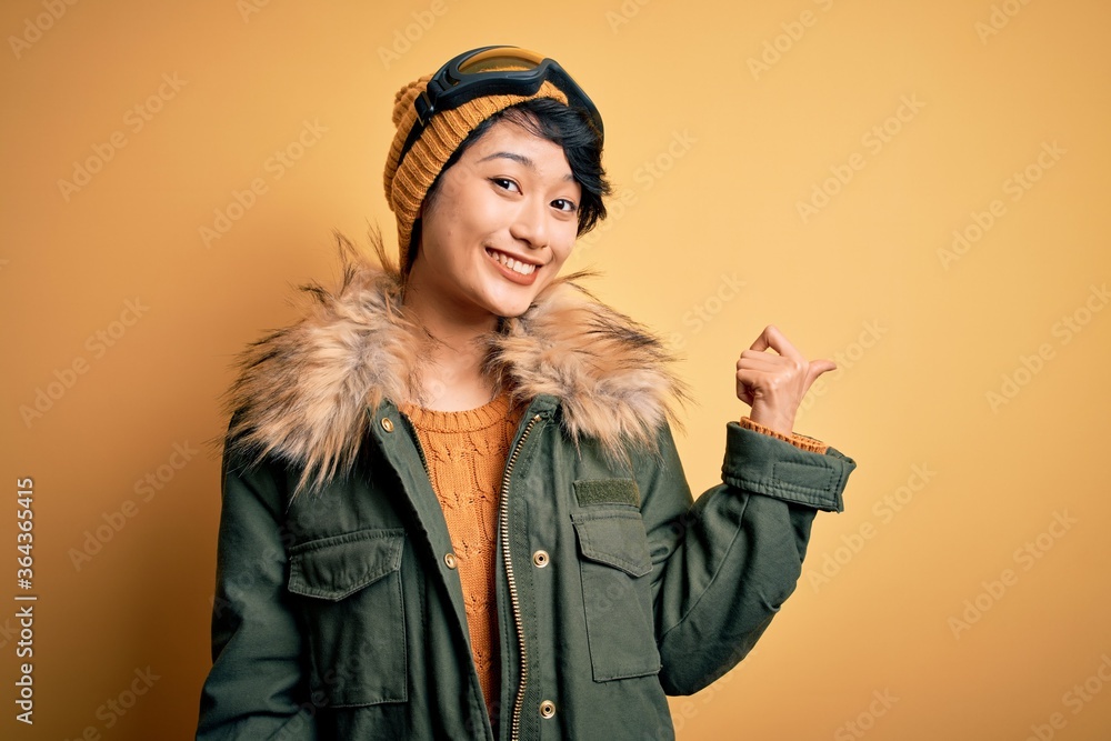 Beautiful asian skier girl wearing snow sportswear using ski goggles over yellow background smiling with happy face looking and pointing to the side with thumb up. Beautiful asian skier girl wearing snow sportswear using ski goggles over yellow background smiling with happy face looking and pointing to the side with thumb up.