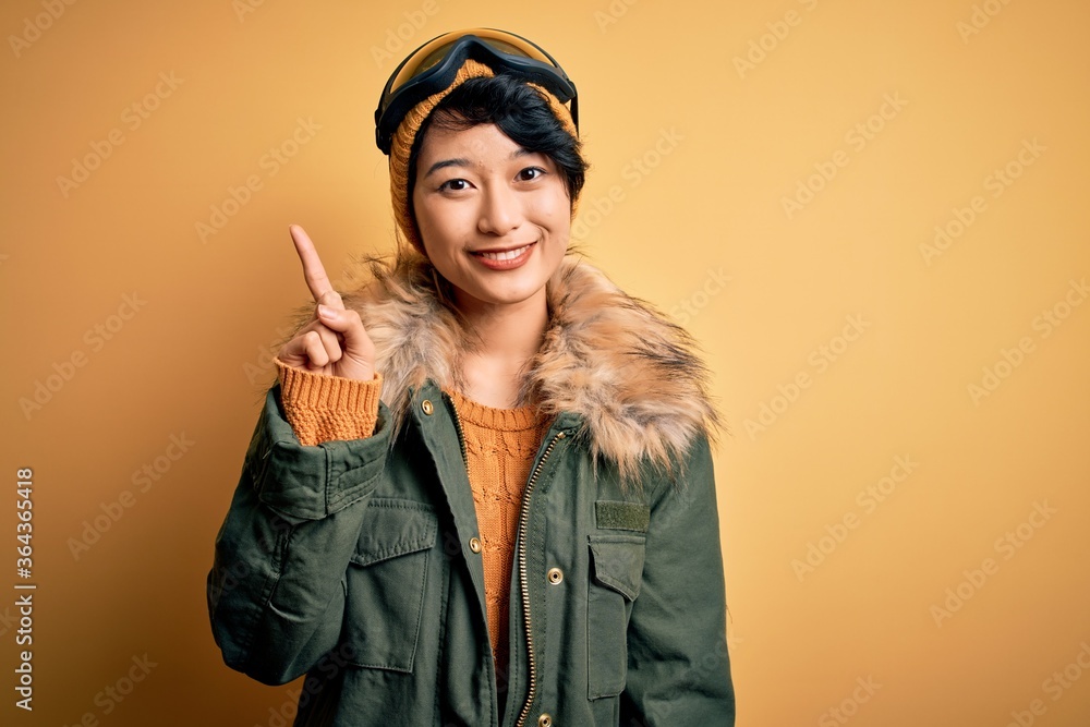 Beautiful asian skier girl wearing snow sportswear using ski goggles over yellow background showing and pointing up with finger number one while smiling confident and happy. Beautiful asian skier girl wearing snow sportswear using ski goggles over yellow background showing and pointing up with finger number one while smiling confident and happy.