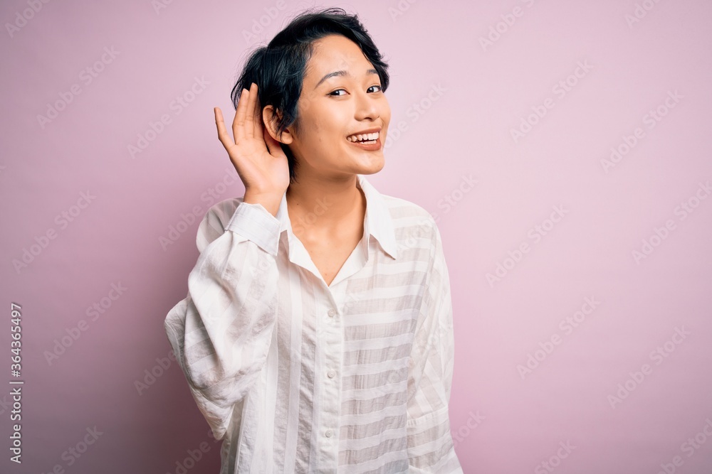Young beautiful asian girl wearing casual shirt standing over isolated pink background smiling with hand over ear listening an hearing to rumor or gossip. Deafness concept. Young beautiful asian girl wearing casual shirt standing over isolated pink background smiling with hand over ear listening an hearing to rumor or gossip. Deafness concept.