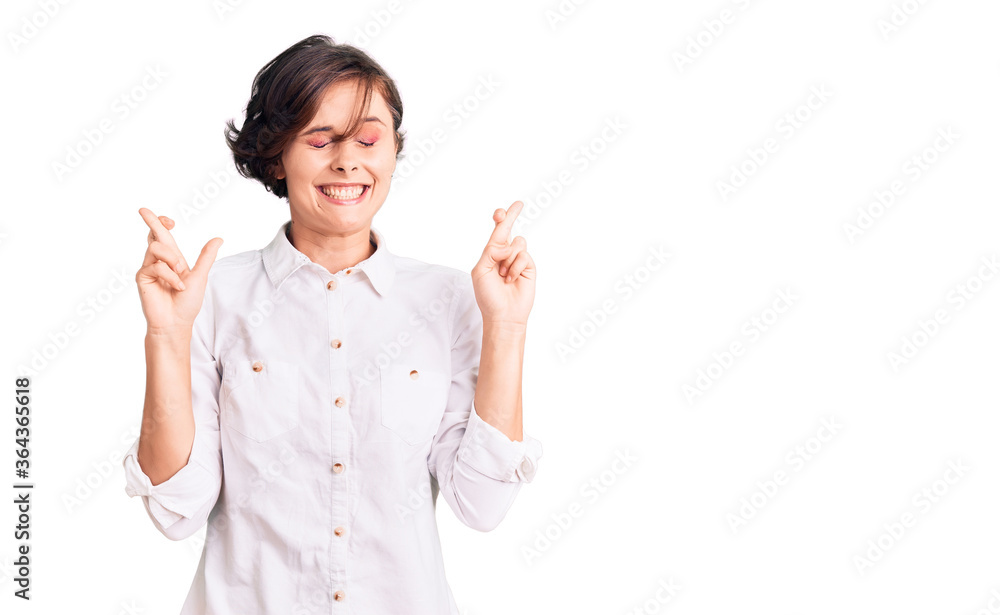 Beautiful young woman with short hair wearing elegant white shirt gesturing finger crossed smiling with hope and eyes closed. luck and superstitious concept. Beautiful young woman with short hair wearing elegant white shirt gesturing finger crossed smiling with hope and eyes closed. luck and superstitious concept.